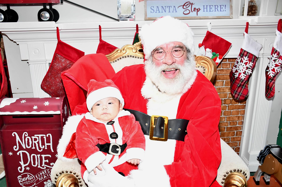 santa seated in front a fireplace with stockings. he is holding an infant wearing a santa suit.